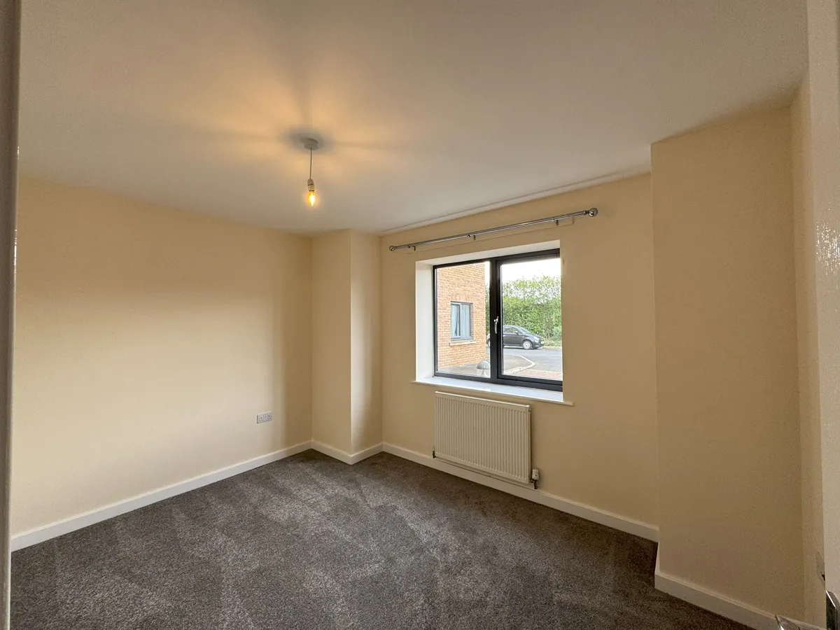 A bedroom with beige walls, grey carpet, window with curtain pole, and white radiator beneath.