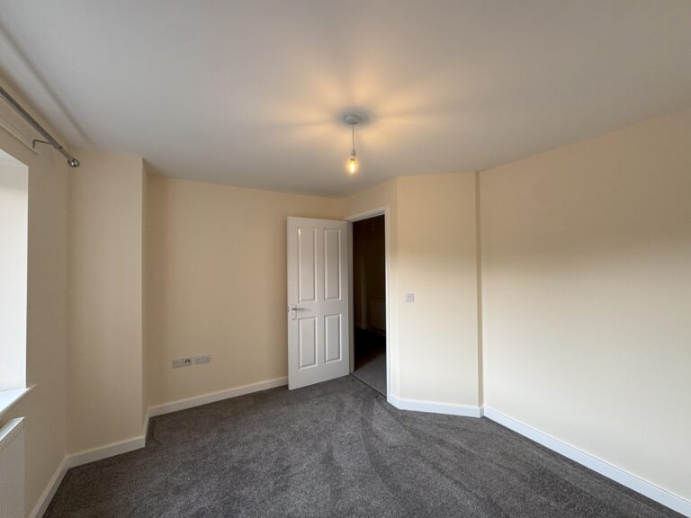 A bedroom with beige walls, grey carpet, window with curtain pole, and a partially open white door leading to a hallway.