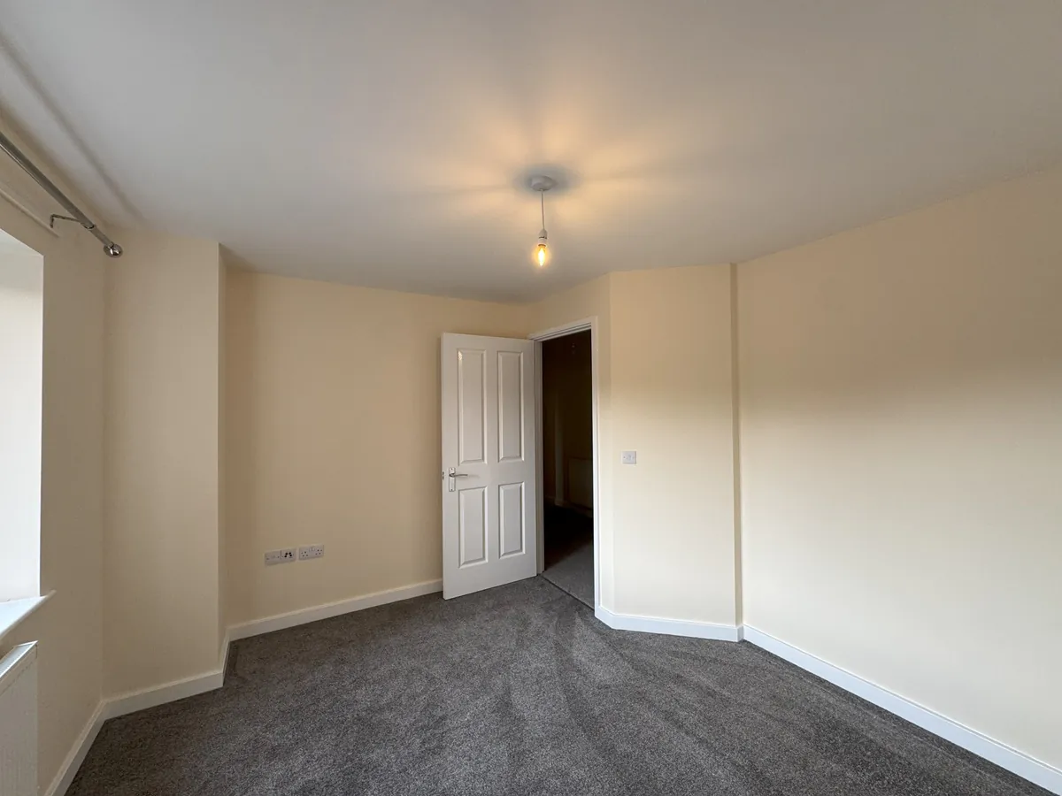 A bedroom with beige walls, grey carpet, window with curtain pole, and a partially open white door leading to a hallway.