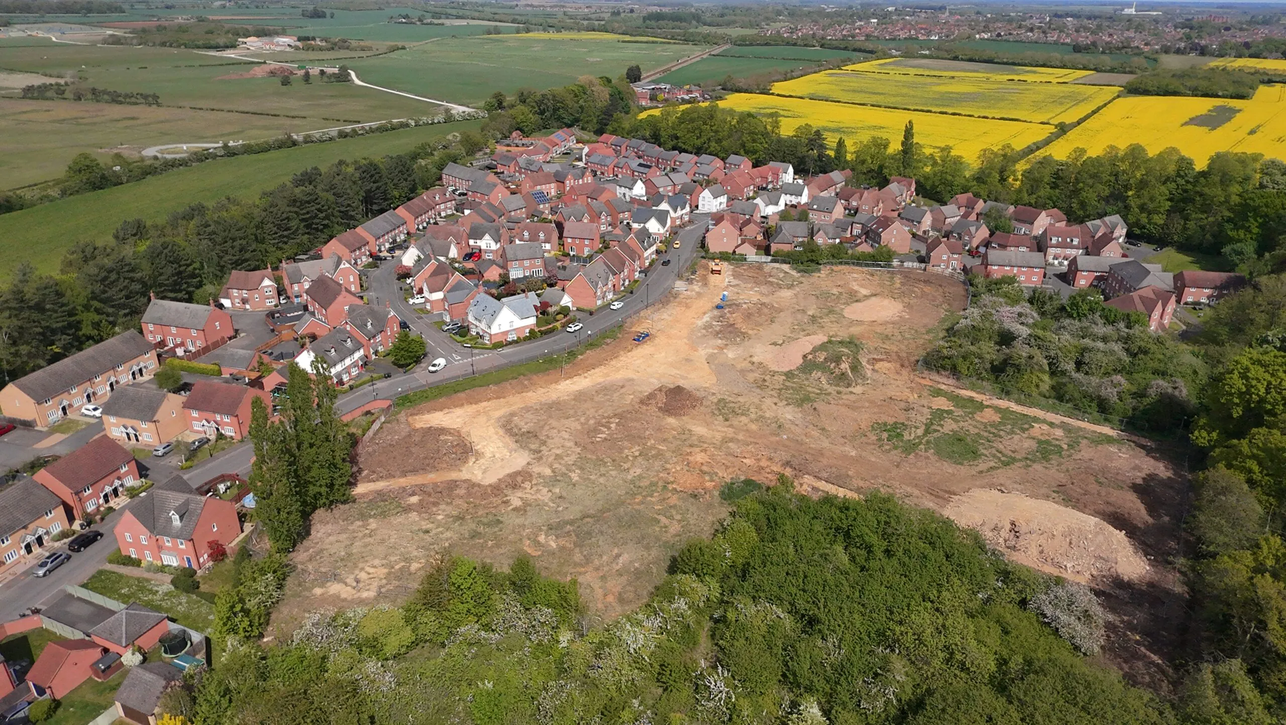 Aerial view of The Orchards at Greylees near Sleaford showing the cleared development site beside existing homes, with surrounding countryside and yellow rapeseed fields in the distance | Lafford Group
