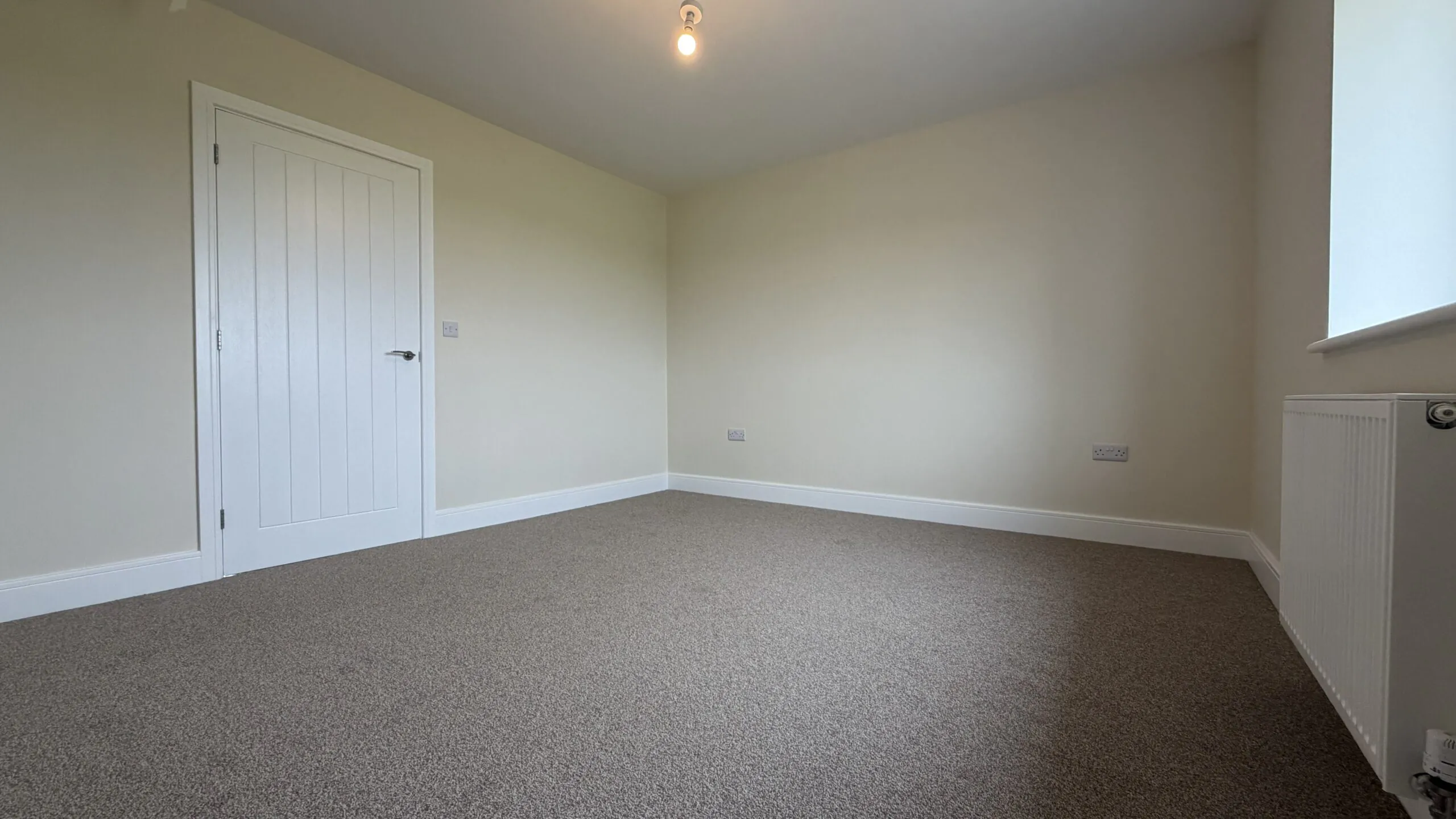 The Waltham at Petrella Place – Bedroom 1 alternate view showing neutral décor, fitted carpet, white internal door and a window with radiator alongside.