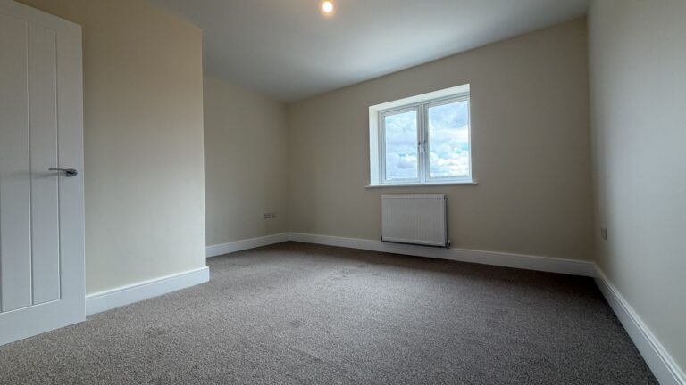 The Waltham at Petrella Place – Bedroom 2 alternate view showing neutral décor, fitted carpet, double-glazed window with radiator beneath, and a partial dividing wall/alcove near the doorway.