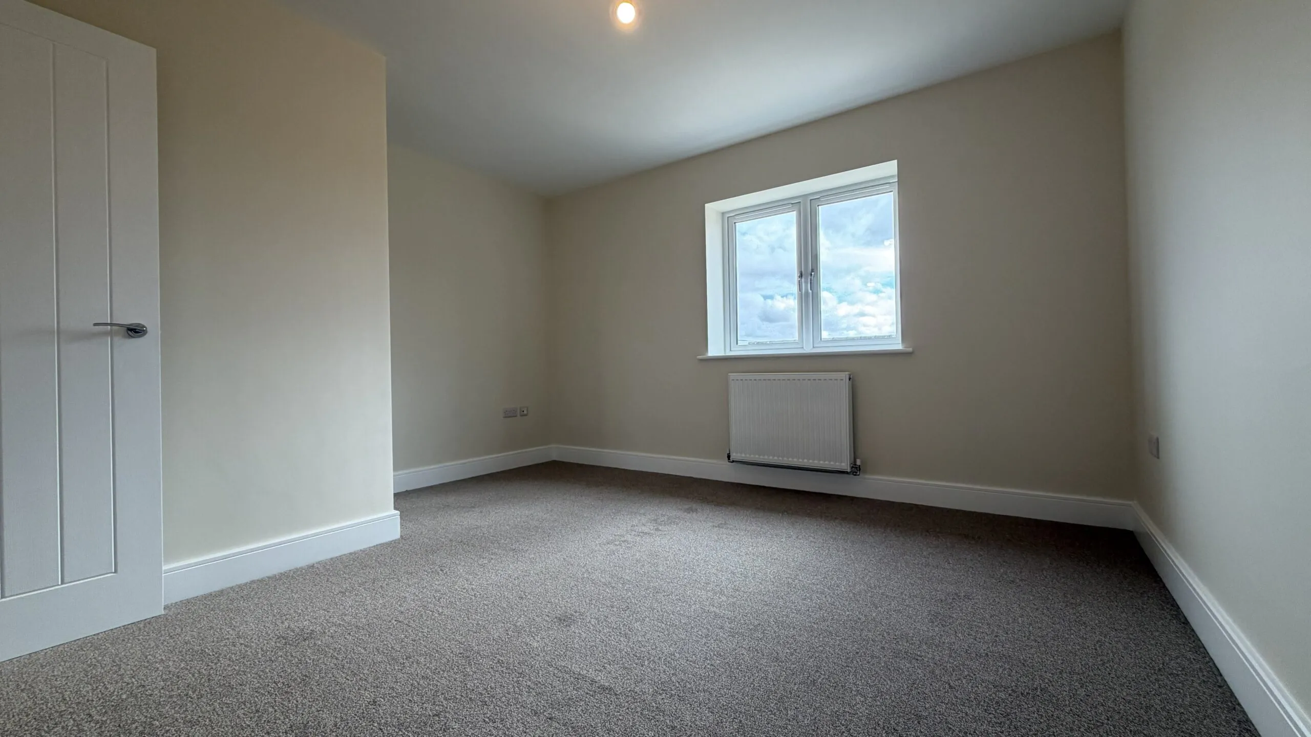 The Waltham at Petrella Place – Bedroom 2 alternate view showing neutral décor, fitted carpet, double-glazed window with radiator beneath, and a partial dividing wall/alcove near the doorway.