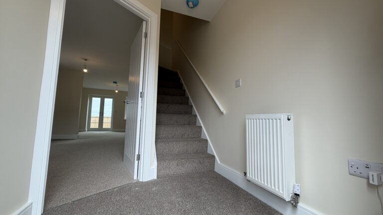 The Waltham at Petrella Place – entrance hallway with carpeted staircase, white handrail, radiator and doorway through to the open-plan living space with French doors beyond.