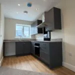 The Waltham at Petrella Place – modern kitchen with dark shaker units, wood-effect worktops, integrated oven and hob, extractor hood and sink beneath a rear window.