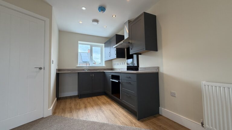 The Waltham at Petrella Place – modern kitchen with dark shaker units, wood-effect worktops, integrated oven and hob, extractor hood and sink beneath a rear window.