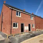 External street view at Petrella Place showing The Waltham Plot 19 on the left and Plot 20 on the right, with Plots 21 and 22 visible further along the row of new red-brick homes.