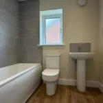 The Caister at Petrella Place – family bathroom with bath and grey tiled surround, toilet below a frosted window, pedestal basin, radiator and wood-effect flooring.