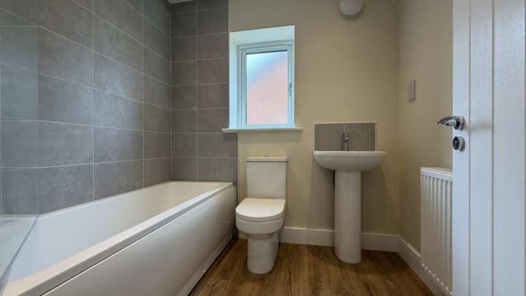 The Caister at Petrella Place – family bathroom with bath and grey tiled surround, toilet below a frosted window, pedestal basin, radiator and wood-effect flooring.