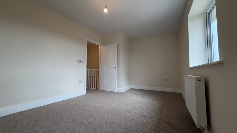 The Caister at Petrella Place – Bedroom 1 alternate view showing neutral décor, fitted carpet, large window with radiator below and the open door to the landing.