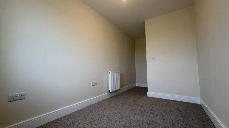 The Caister at Petrella Place – Bedroom 3 alternate view showing neutral décor, fitted carpet, a wall-mounted radiator and a partial dividing wall creating a defined entrance area.