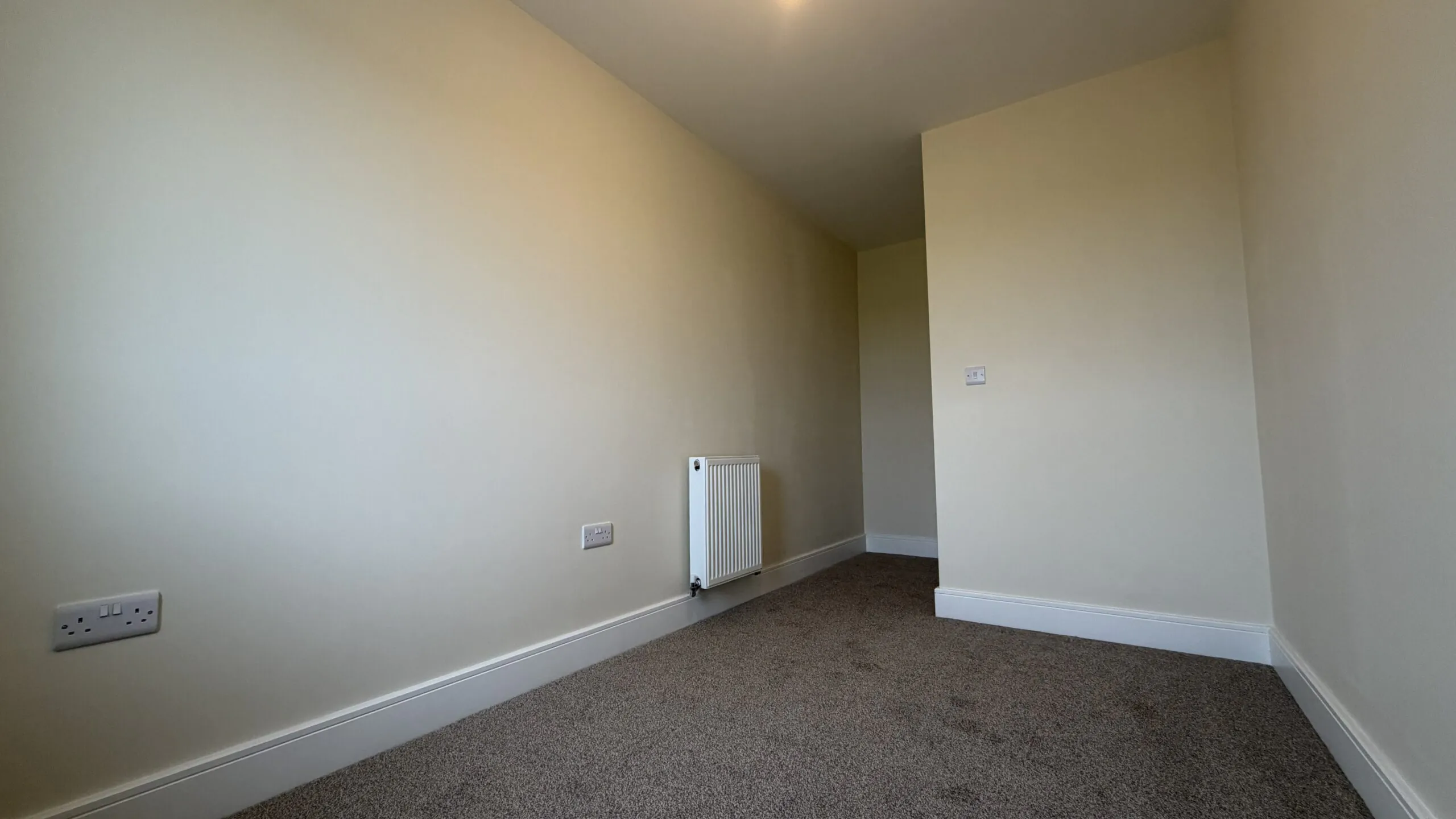 The Caister at Petrella Place – Bedroom 3 alternate view showing neutral décor, fitted carpet, a wall-mounted radiator and a partial dividing wall creating a defined entrance area.