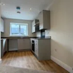 The Caister at Petrella Place – modern kitchen with grey units, wood-effect worktops, integrated oven and hob, sink under a rear window and recessed ceiling spotlights.