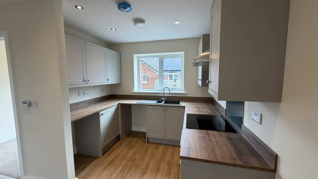 The Caister at Petrella Place – close-up of the kitchen showing grey units, wood-effect worktops, sink under a window and an electric hob with extractor above.