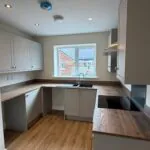 The Caister at Petrella Place – close-up of the kitchen showing grey units, wood-effect worktops, sink under a window and an electric hob with extractor above.