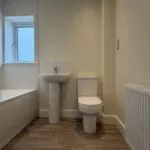 The Caister at Petrella Place – family bathroom with bath and tiled surround, pedestal basin, toilet, window and wood-effect flooring.