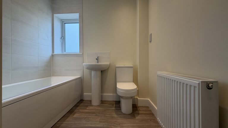 The Caister at Petrella Place – family bathroom with bath and tiled surround, pedestal basin, toilet, window and wood-effect flooring.