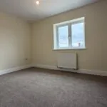 The Caister at Petrella Place – Bedroom 1 with neutral décor, carpeted floor, large window and radiator beneath.