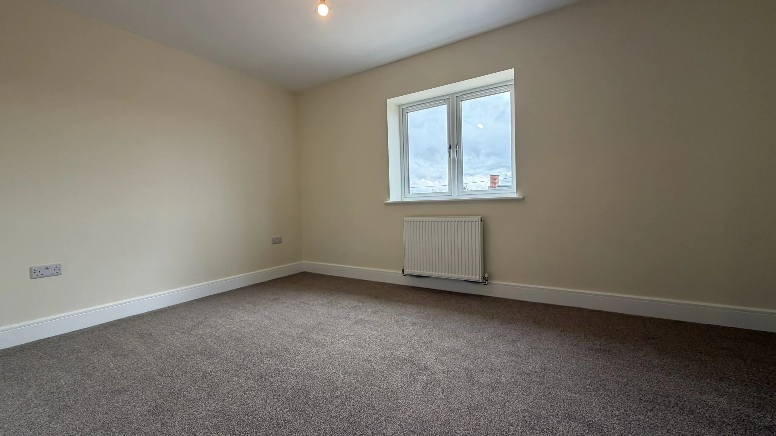 The Caister at Petrella Place – Bedroom 1 with neutral décor, carpeted floor, large window and radiator beneath.