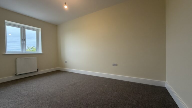 The Caister at Petrella Place – Bedroom 2 with neutral décor, carpeted flooring, window and radiator beneath.