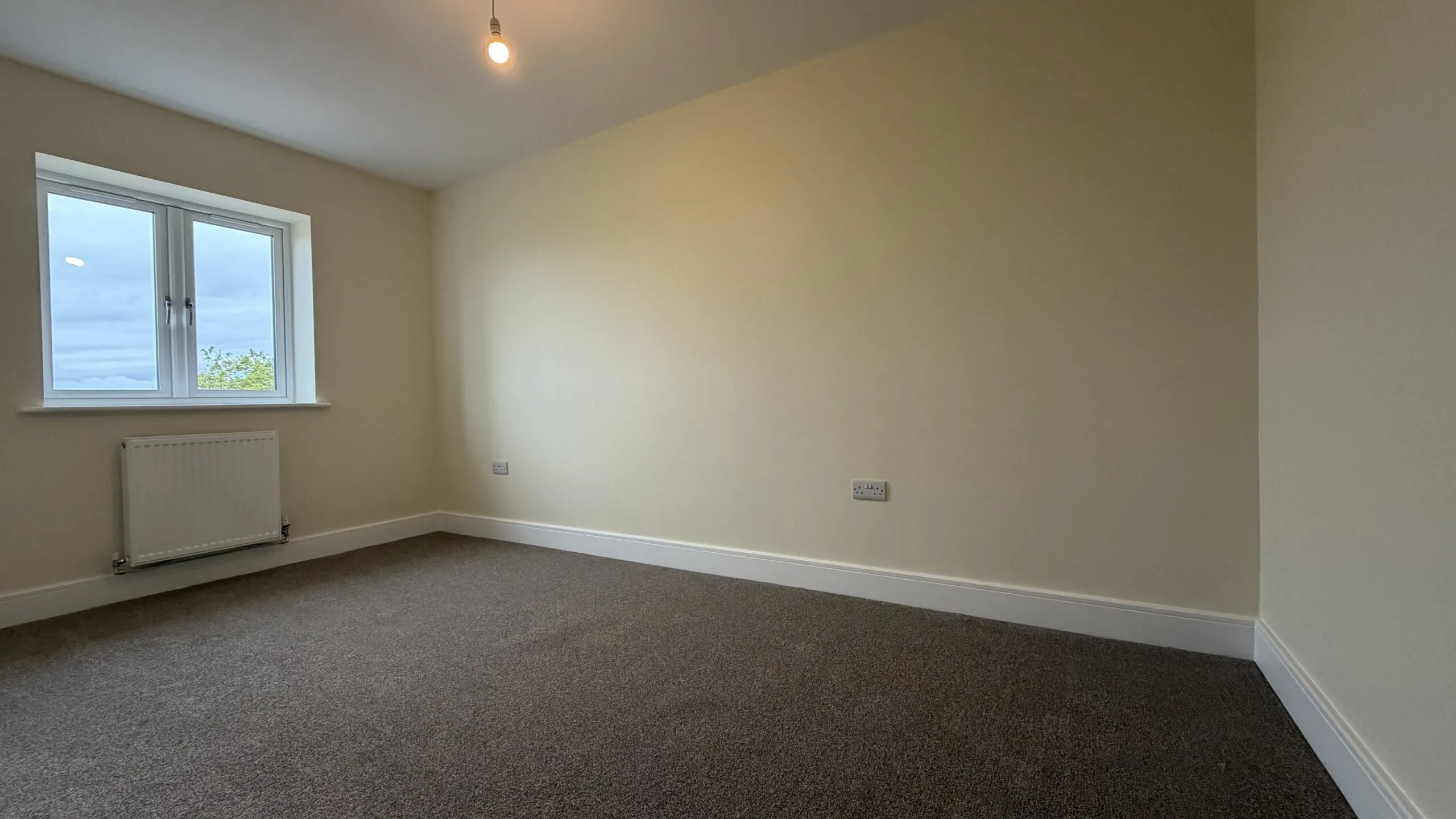 The Caister at Petrella Place – Bedroom 2 with neutral décor, carpeted flooring, window and radiator beneath.
