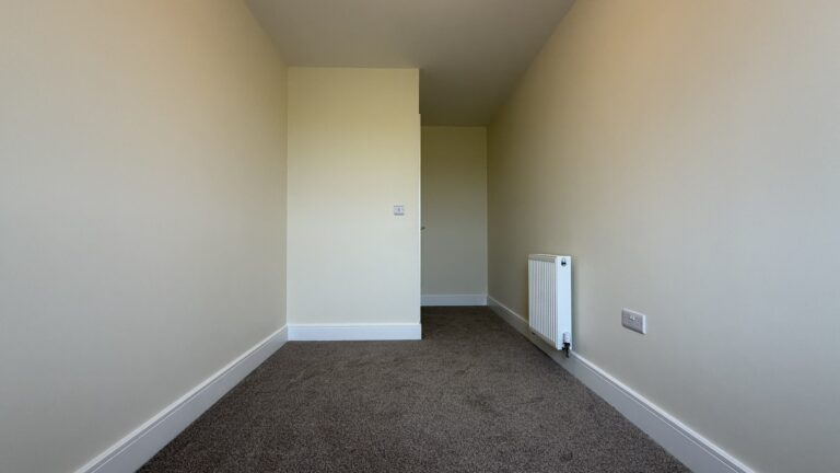 The Caister at Petrella Place – Bedroom 3 alternate view showing a long, narrow room with neutral décor, fitted carpet, radiator and the doorway area.