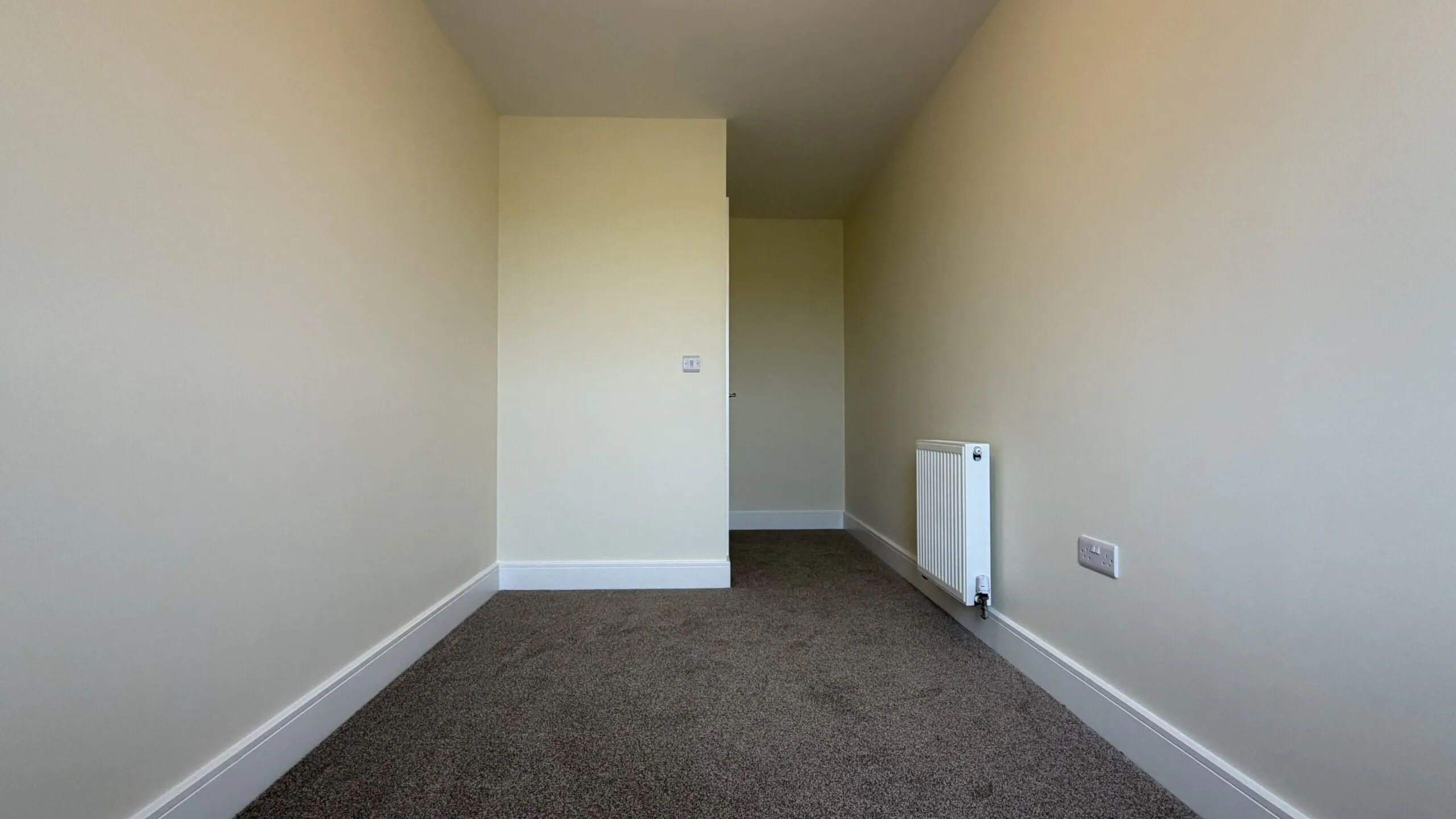 The Caister at Petrella Place – Bedroom 3 alternate view showing a long, narrow room with neutral décor, fitted carpet, radiator and the doorway area.