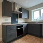 The Caister at Petrella Place – modern U-shaped kitchen with dark cabinetry, wood-effect worktops, integrated oven, stainless extractor and sink beneath a bright window.