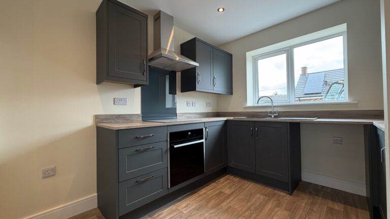 The Caister at Petrella Place – modern U-shaped kitchen with dark cabinetry, wood-effect worktops, integrated oven, stainless extractor and sink beneath a bright window.
