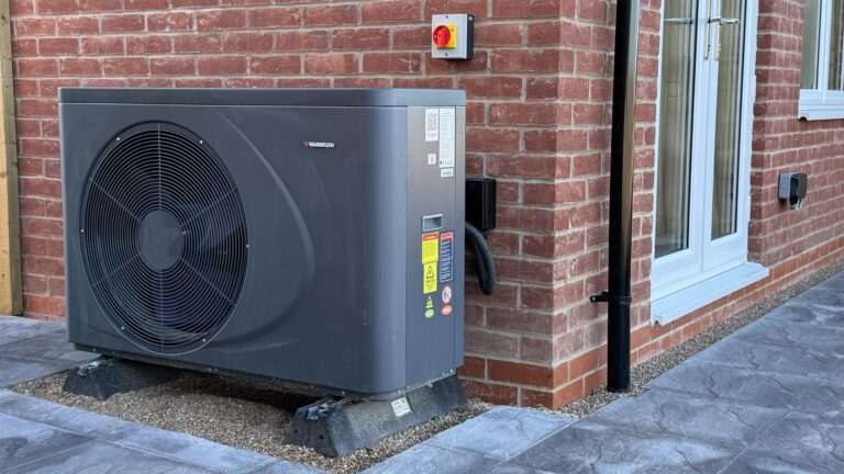 Warmflow air source heat pump external unit beside a red brick wall at Petrella Place, with an isolator switch above and a window nearby.
