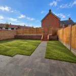 Enclosed rear garden at 34 Warwick Drive featuring a paved patio, lawned garden, secure timber fencing and raised rear section with steps providing additional outdoor space.